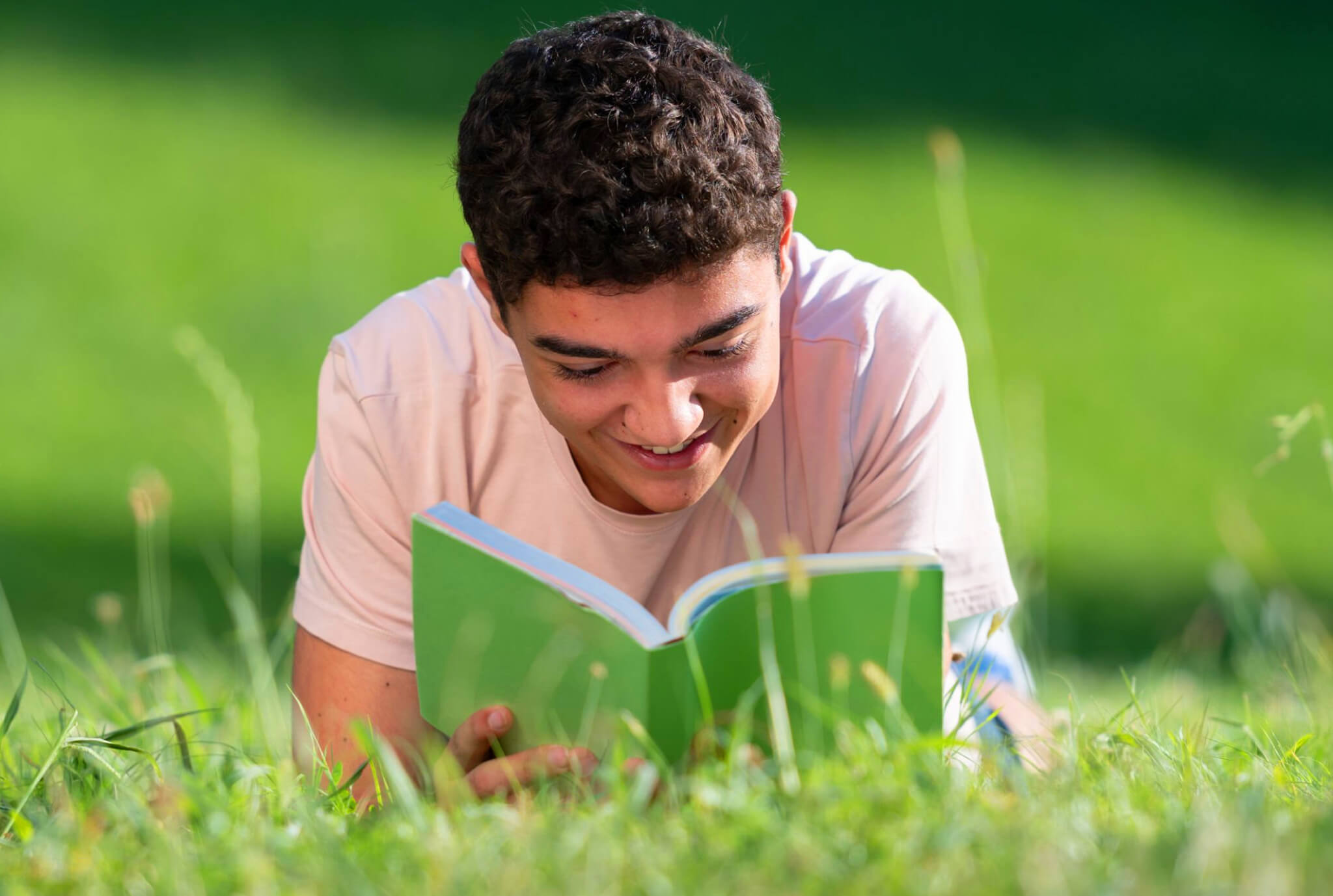 A smiling boy lays in the grass reading a book